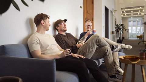Tre mænd sidder på en sofa i et lyst lokale, griner og drikker kaffe, med planter og en træbord i baggrunden.